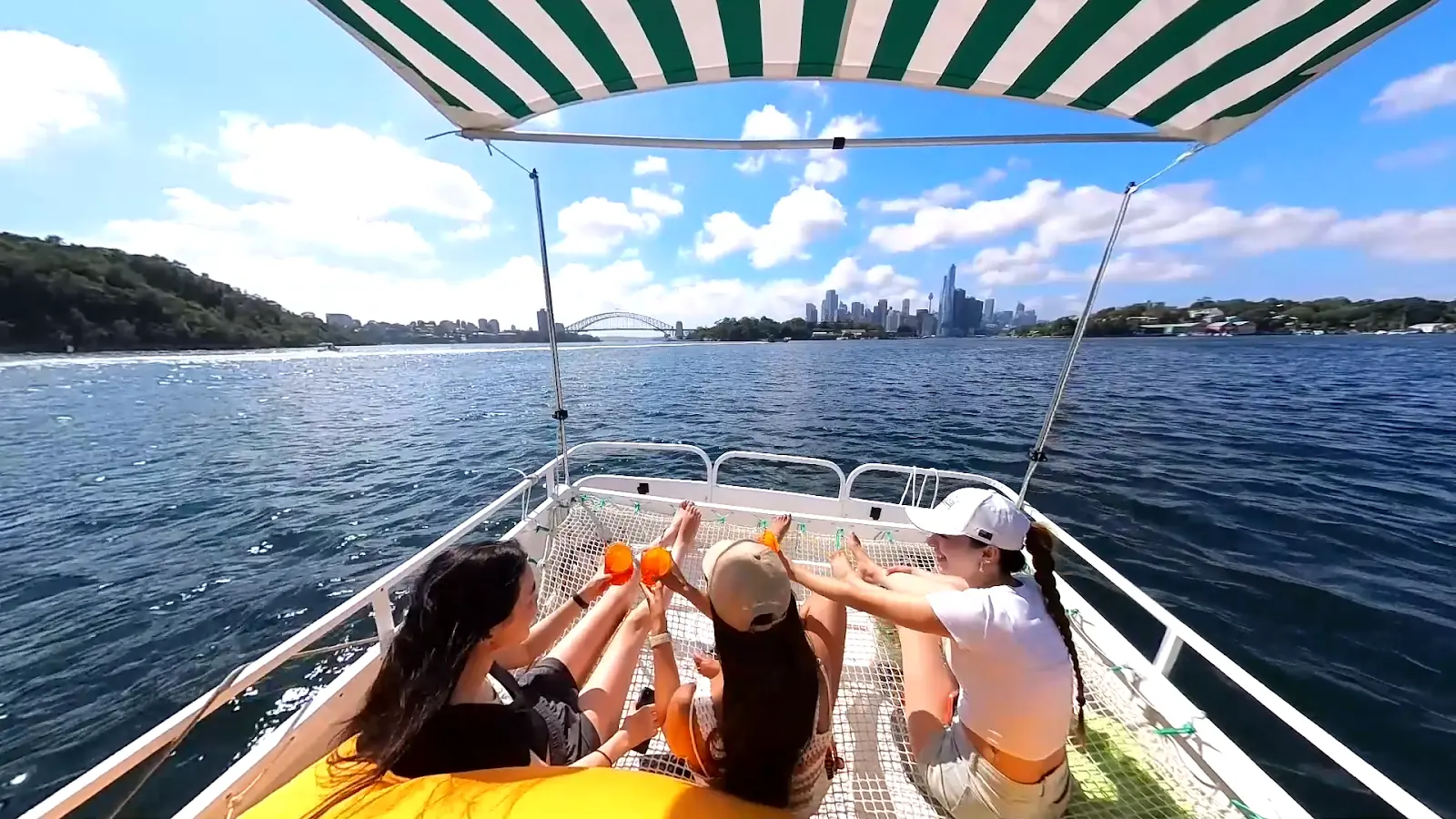 Three friends on Capri's trampoline sunbed raising Aperol cups, the city skyline and Harbour Bridge in the distance